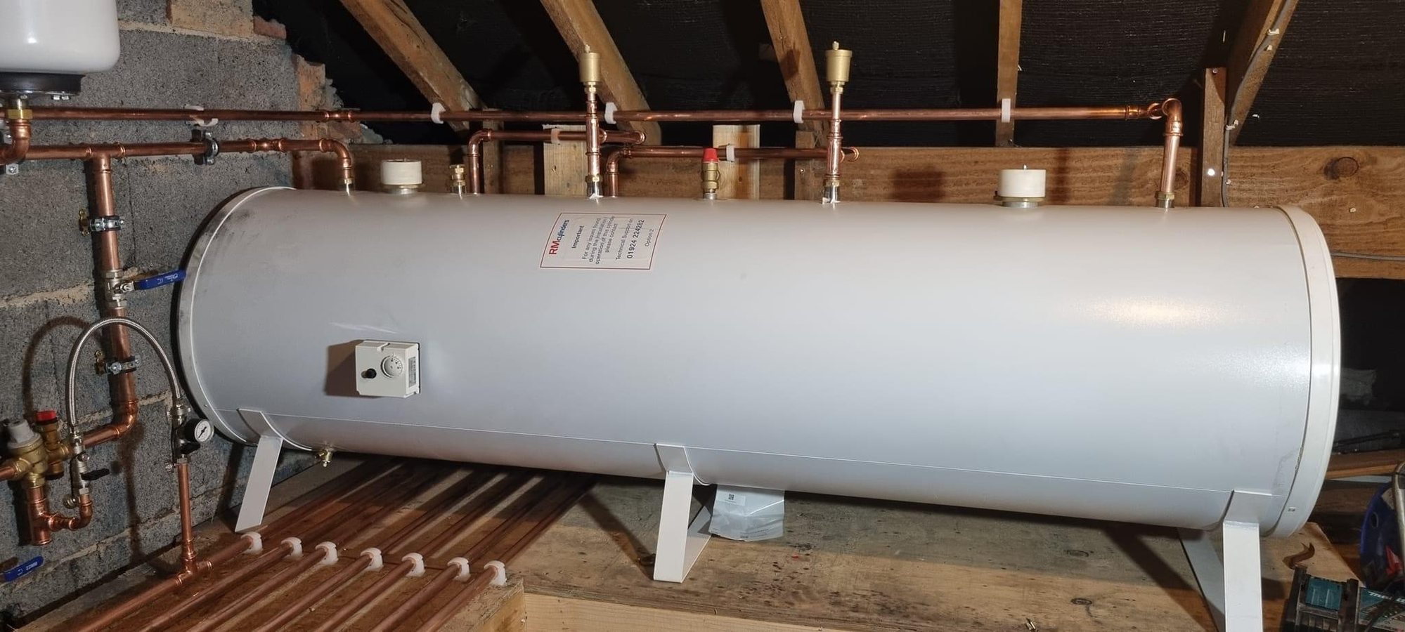 Horizontal unvented cylinder set on brackets in the loft, with the safety group, expansion vessel and tundish all accessible.